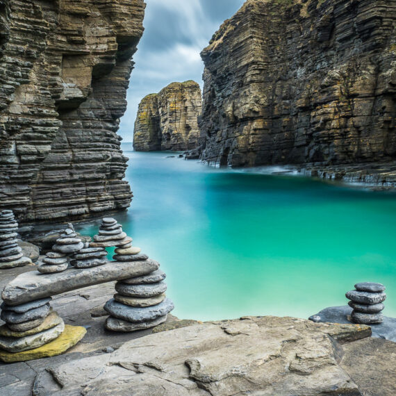 Travel photography Scotland: Cairns formed from stacks of pebbles beside turquoise seawater in a natural harbour below Sinclair Girnigoe Castle near Wick, Scotland.