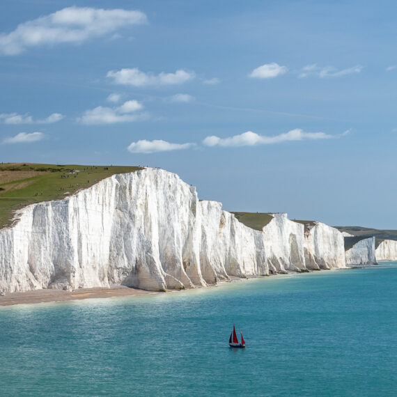 Travel photography England: A yacht with red sails seen in the English Channel by the Seven Sisters, chalk cliffs in East Sussex, England.