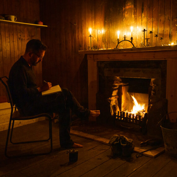 A writer sits barefoot in front of an open fire in a bothy, with his boots and socks by the fire and a hip flask by his side; the scene is lit entirely by the fire and a few candles