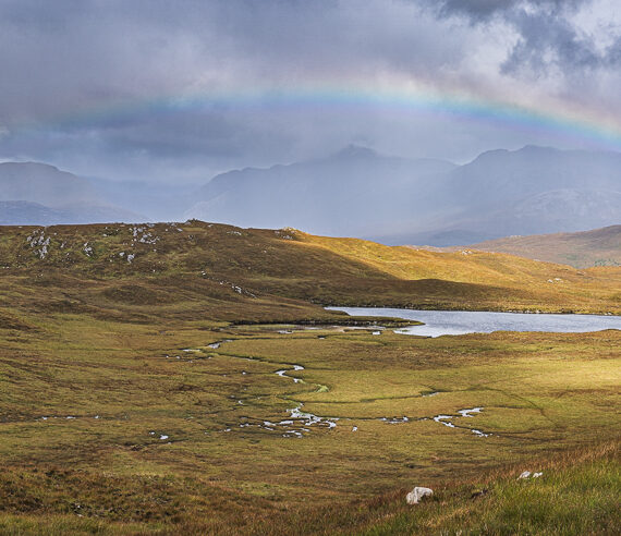 A rainbow spans a lake and moor in front of a mountain range under dark, cloudy skies in Torridon, Scotland
