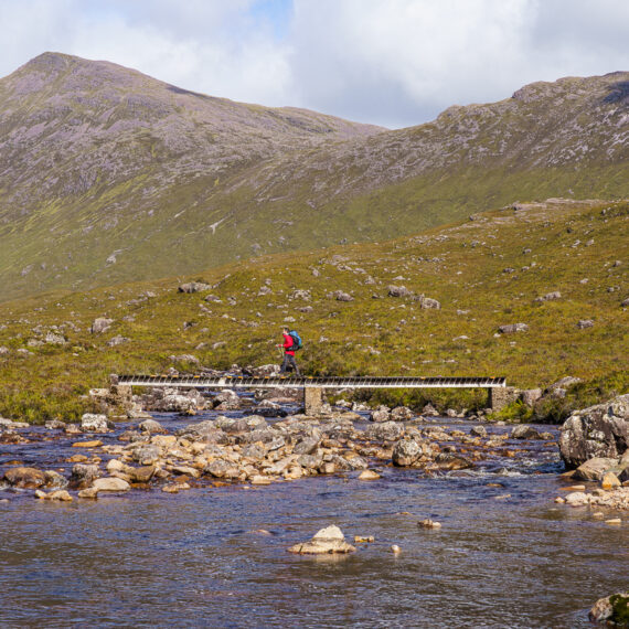 A hiker in a red coat crosses a wooden footbridge over a river strewn with bolders, in front of bare peaks in Coire Fionnaraich, Scotland