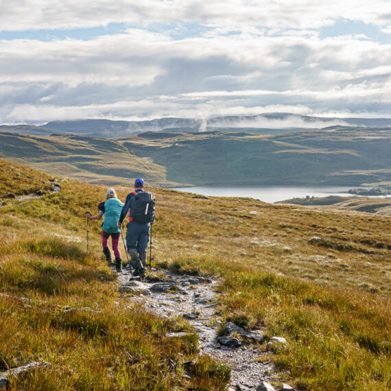 A male and female hiker walk along a path towards a cloudy and hilly landscape in Fisherfield, Scotland
