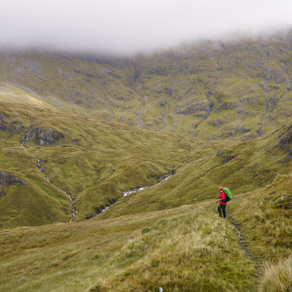 A male hiker in a red coat looks out at a high mountain landscape, fully enclosed by green slopes, streams and clouds, on Ben More Assynt in Scotland