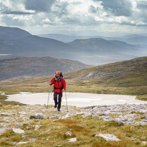 A male hiker in a red coat hiking uphill from Inchnadamph in Assynt with stormy skies and mountains stretching out into the horizon